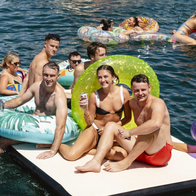 A group of people enjoying a sunny day on the water. They are sitting on inflatable loungers and a platform. One person holds a drink. A boat is in the background, and everyone appears relaxed and happy.