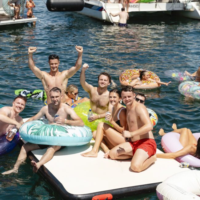 A group of people enjoying a sunny day on the water. Some are sitting on inflatable mats and tubes, raising their arms and holding drinks. Boats are visible in the background. The mood is festive, with the water sparkling under the sun.