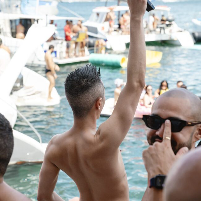 A person in yellow swimwear, seen from behind, raises a colorful fan high. They stand by the water where boats and people are visible in the background. The scene is lively, with others nearby enjoying the sunny day.