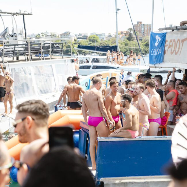 A vibrant boat party with people wearing colorful swimwear, mostly pink. The deck is crowded with partygoers socializing and enjoying the sunny weather. Lifebuoys and banners are visible on the boat, and a distant shoreline is in the background.