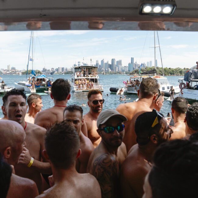 A large group of people in swimsuits gather on a boat, chatting and enjoying the sunny day. Other boats are visible, and a city skyline is in the background under a clear blue sky.