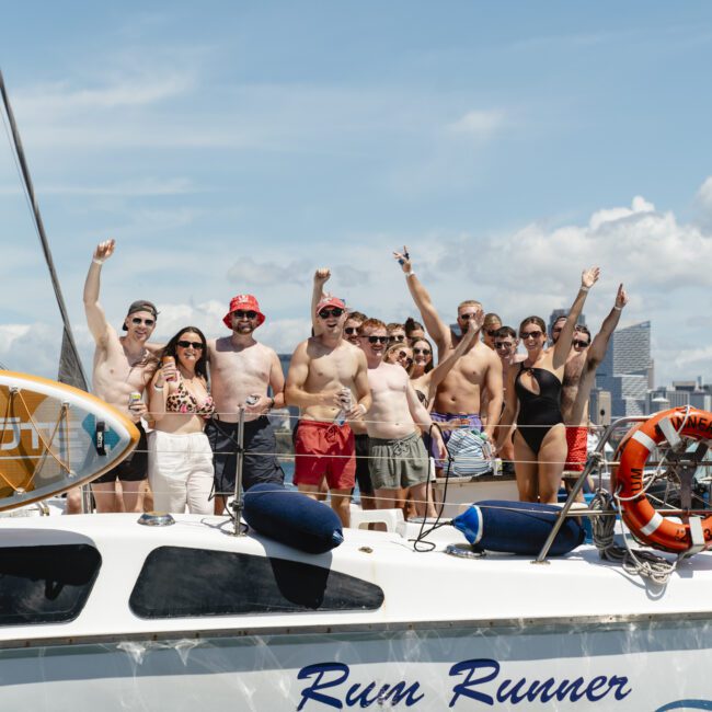 A group of people in swimwear stand on a boat named "Rum Runner," posing happily with arms raised. The boat is on water with a city skyline in the background under a cloudy sky.