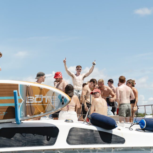 A group of people enjoy sunny weather on a boat. Some are standing and talking, while one person raises their arms in excitement. A paddleboard is seen on the boat's deck. The boat's name, partially visible, reads "Rum R...".
