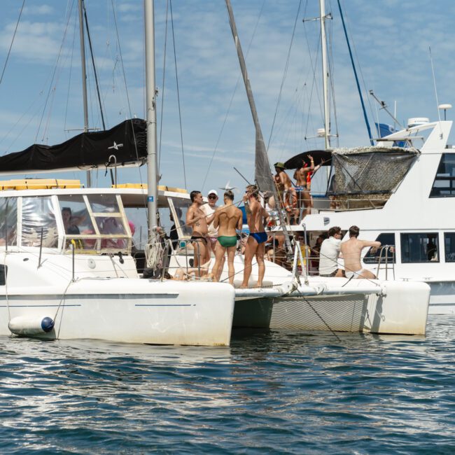 A group of people in swimwear stand and sit on two docked boats in the water. One boat is a catamaran with a colorful flag, and the other is a larger yacht. The sky is partly cloudy.