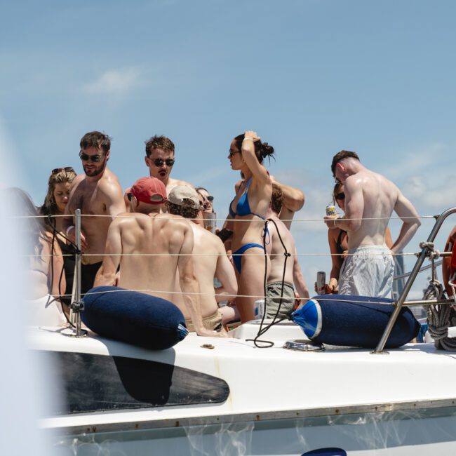 A group of people in swimsuits are gathered on a boat under a clear blue sky. They are chatting and enjoying the sunny day. A life preserver is visible on the railing of the boat.