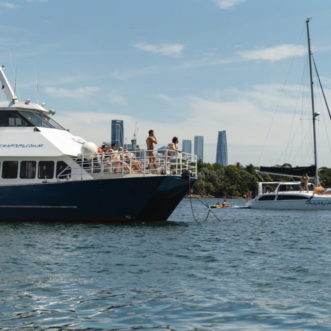 A boat with people on its deck is anchored near the shore with city skyscrapers in the background. A smaller sailboat is nearby, and another boat is partially visible. The sky is clear, and the water is calm.