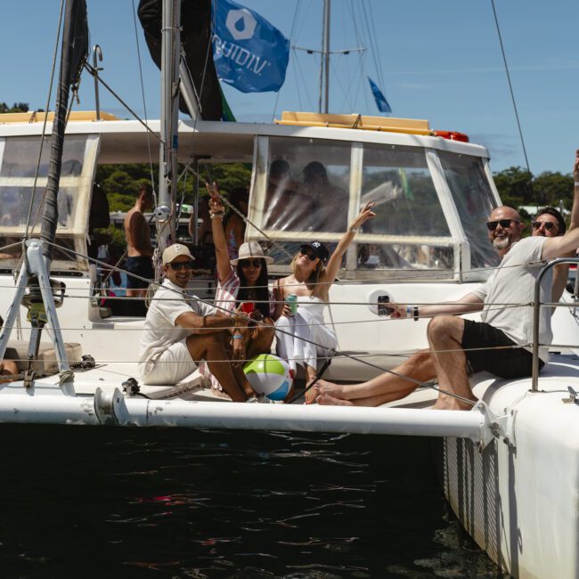 A group of people is sitting and standing on the deck of a catamaran, smiling and waving their arms. They appear to be enjoying a sunny day on the water. A beach ball is on the deck, and other boats are visible in the background.