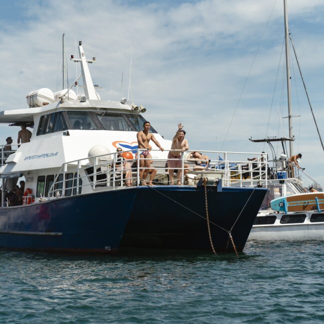 A large blue and white boat on the water with people standing on its deck, enjoying a sunny day. Nearby, smaller boats are visible against a blue sky with scattered clouds.