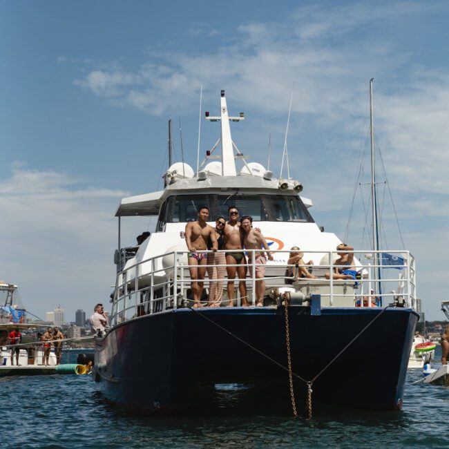 A large boat with several people on the deck floats on the water. Three men in swimwear stand at the front, and others are seated or standing around. The sky is partly cloudy, and there are buildings visible in the distant background.