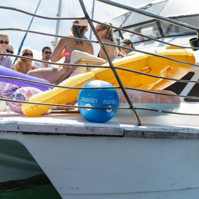 People relaxing on a catamaran, with colorful inflatable airbeds and a blue ball on the deck. The sky is clear, and several individuals are sitting on the boat, enjoying the sunny weather.