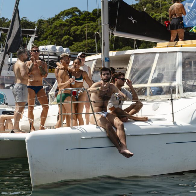 A group of people in swimwear relax on the deck of a catamaran under sunny skies. Some are sitting or lying down, while others stand, holding drinks. Surrounding them are lush green trees and other docked boats.