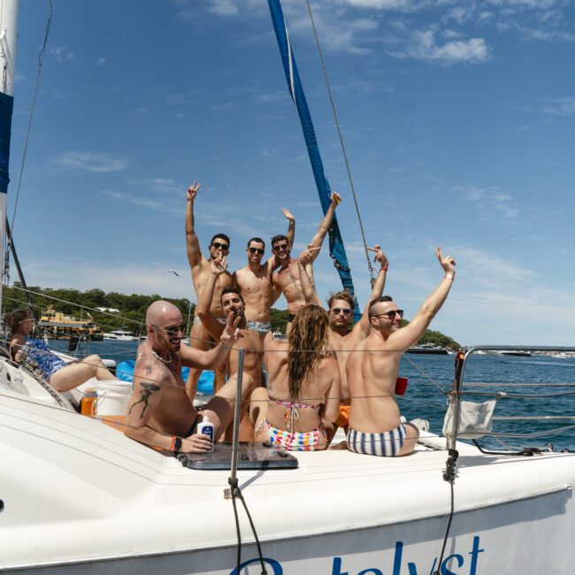 A group of people in swimsuits are posing and smiling on a sailboat named "Catalyst." They are raising their arms and making peace signs. The boat is docked by the water under a clear blue sky.