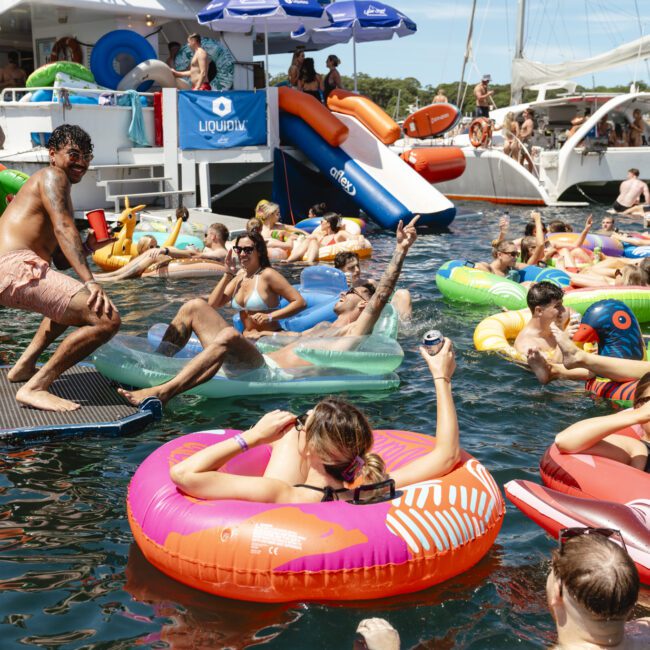 A lively pool party scene with people relaxing on colorful inflatable rings in the water, holding drinks. A boat with a slide is in the background, and a group appears to be enjoying the sunny day and festive atmosphere.