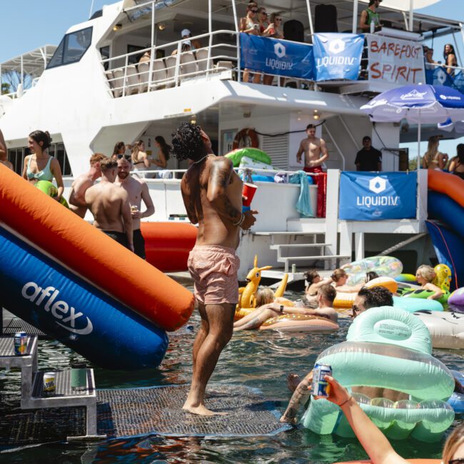 People are enjoying a pool party on a boat, with inflatable toys and slides. The scene is lively with guests swimming and socializing in the water. The boat is adorned with banners, and the sun is shining.