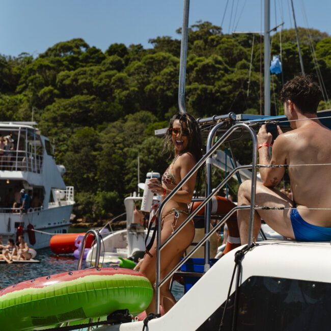 A woman in a bikini stands on a boat holding a drink, smiling. A man sits nearby, wearing swim trunks. There's a large inflatable and another boat in the background, with green hills in the distance. The scene is sunny and festive.