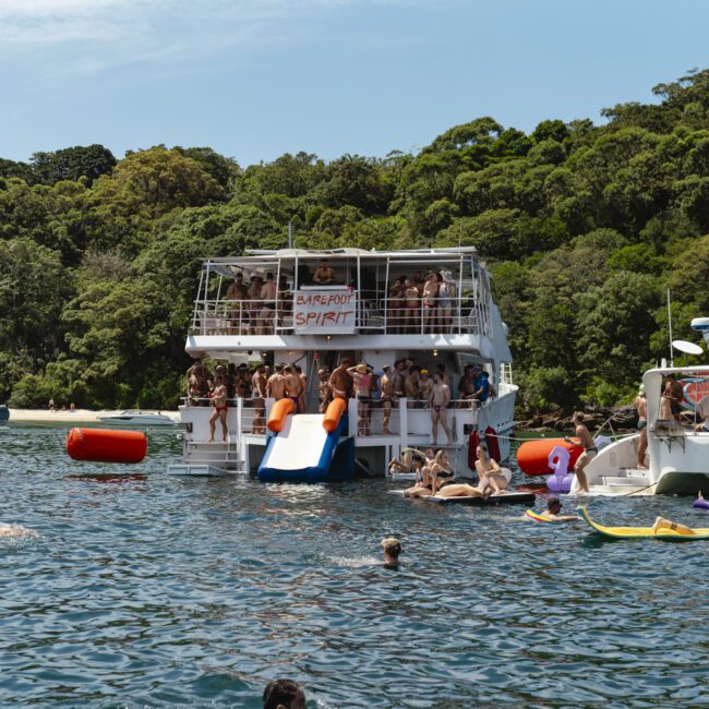 A lively scene on the water with a large boat hosting a party. People enjoy slides and swimming, with others relaxing on smaller boats nearby. The backdrop features lush green trees under a clear blue sky.