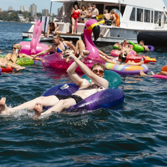 A group of people enjoying a sunny day on a lake, floating on colorful inflatable rings near a boat. The scene includes a giant pink flamingo float, with partygoers relaxed and socializing in swimsuits.