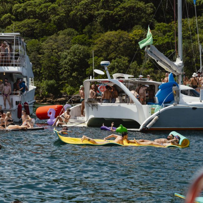 People enjoying a sunny day on a lake, with multiple boats docked nearby. Some are lounging on inflatable rafts in the water, while others socialize on the boats. Lush greenery lines the background, indicating a scenic and lively atmosphere.