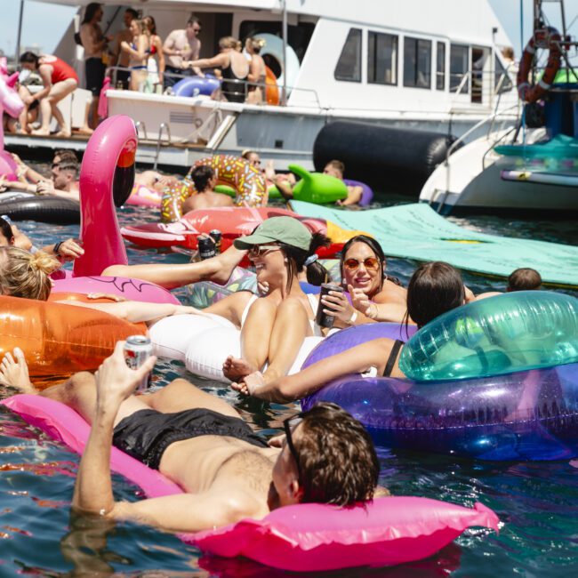 A group of people relax on colorful inflatable floats in the water near boats. They are enjoying sunny weather with drinks in hand. The scene is lively, with an inflatable flamingo and unicorn visible. The water is clear and blue.