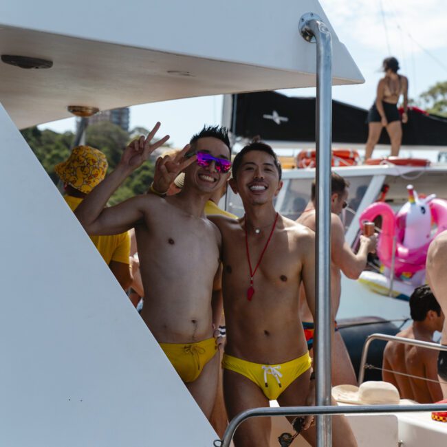 Two people in yellow swimwear stand on a boat, smiling and posing with peace signs. They are surrounded by others in the background, enjoying a sunny day on the water.