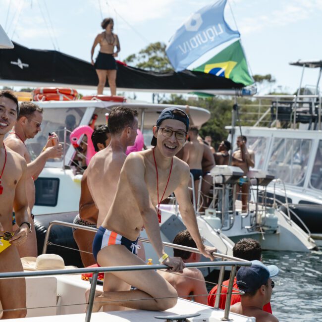 A group of people in swimwear enjoy a sunny day on boats. One person in glasses and a cap kneels on the bow, smiling. Others socialize on nearby boats, with a Brazilian flag visible. The scene conveys a lively, festive atmosphere on the water.