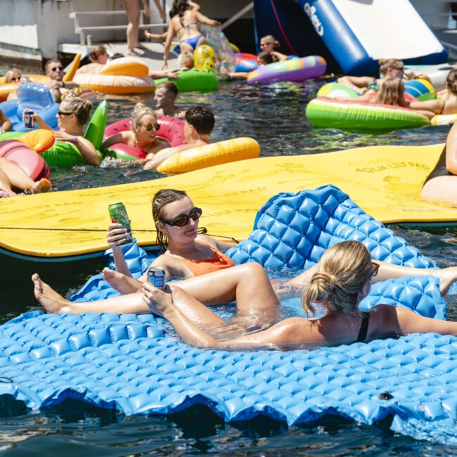 A lively scene of people enjoying a sunny day on the water. Two individuals relax on a blue inflatable mat, holding drinks. Around them, others float on colorful inflatables near a large yellow mat. A boat and a water slide are in the background.