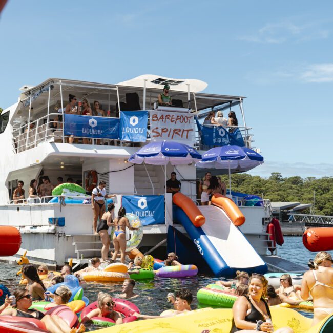 A large boat party is taking place on the water. People are swimming and lounging on colorful inflatables around the boat. The boat has slides and banners that read "BAREFOOT SPIRIT" and "LIQUID." The background features trees and a sunny sky.