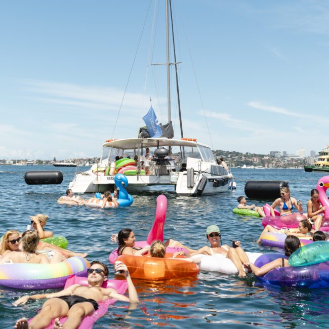 A group of people relax on colorful inflatable rings in the water, surrounded by boats. A catamaran and other vessels are nearby, and the skyline is visible in the distance under a clear blue sky.