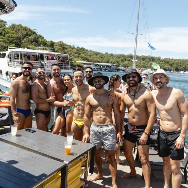 A group of people in swimwear are posing on a boat. They are smiling and standing together under a bright blue sky. Two boats are visible in the background, with trees lining the shore.