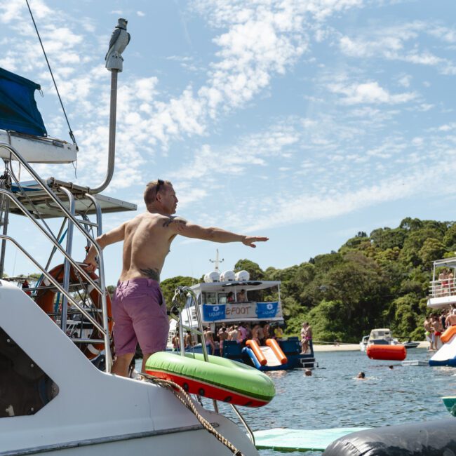 A man in swim trunks stands on the edge of a boat, preparing to jump into the water. Other boats with people and inflatable slides are in the background, near a wooded shoreline under a clear sky.