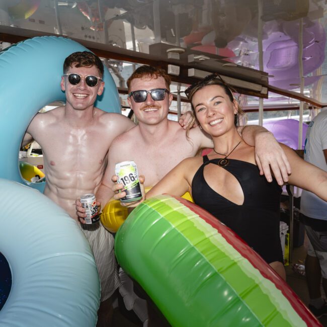 Three people in swimsuits are smiling and holding inflatable rings on a boat. They are wearing sunglasses and holding canned drinks, enjoying a lively atmosphere with others in the background.