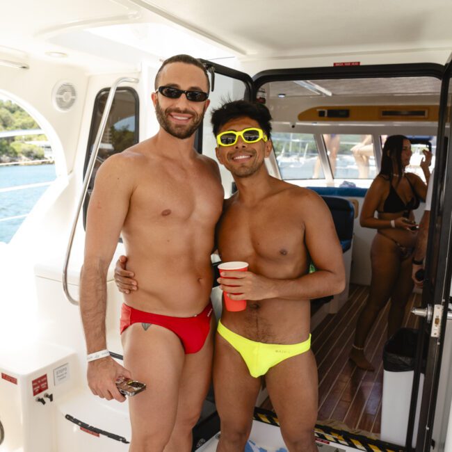 Two people in swimwear pose together on a boat. One wears red trunks, the other yellow trunks and sunglasses. They are smiling and holding drinks. Others are visible in the background on the boat. The scene is sunny and relaxed.