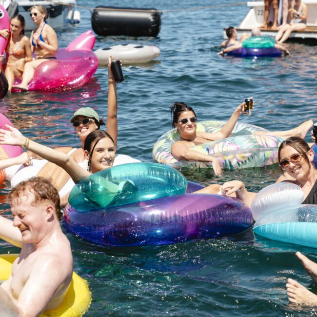 A group of people relaxing on colorful inflatable rings in a lake, holding drinks and smiling. They are surrounded by inflatables shaped like a flamingo and a unicorn. The sun is shining, creating a festive and joyful atmosphere.