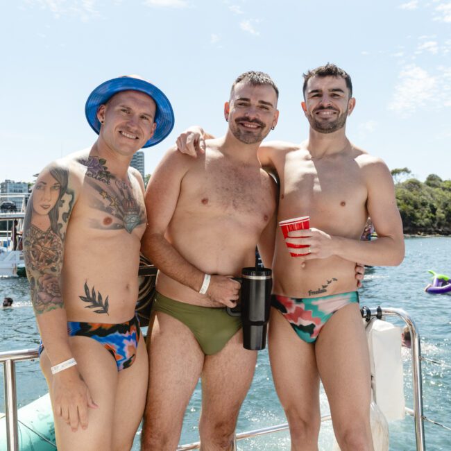 Three men in swimsuits stand on a boat, smiling at the camera. The man on the left wears a bucket hat and has tattoos. They hold drinks, and the background shows water, trees, and inflatable objects. It's a sunny day.