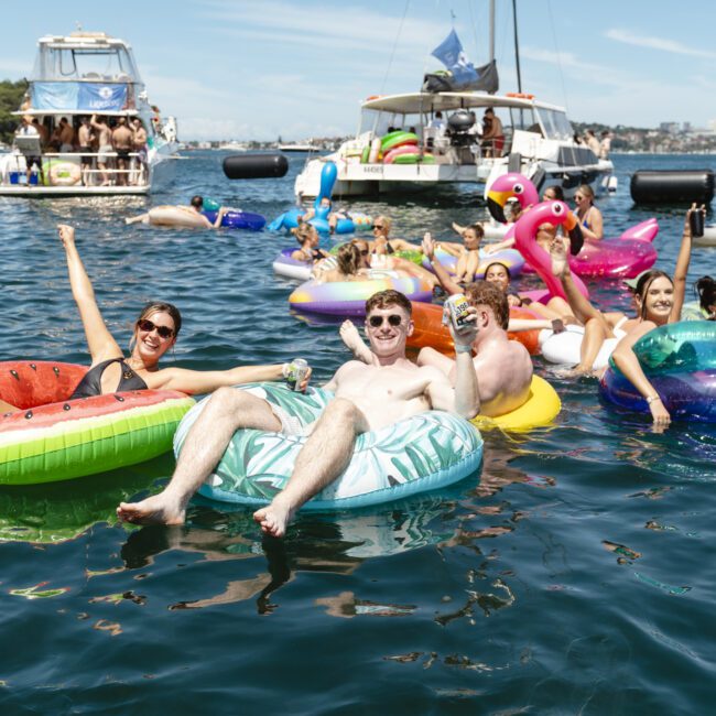 A group of people enjoying a sunny day on the water, lounging on colorful inflatable floats, including a unicorn and a flamingo. Boats are in the background, and everyone looks relaxed and cheerful.