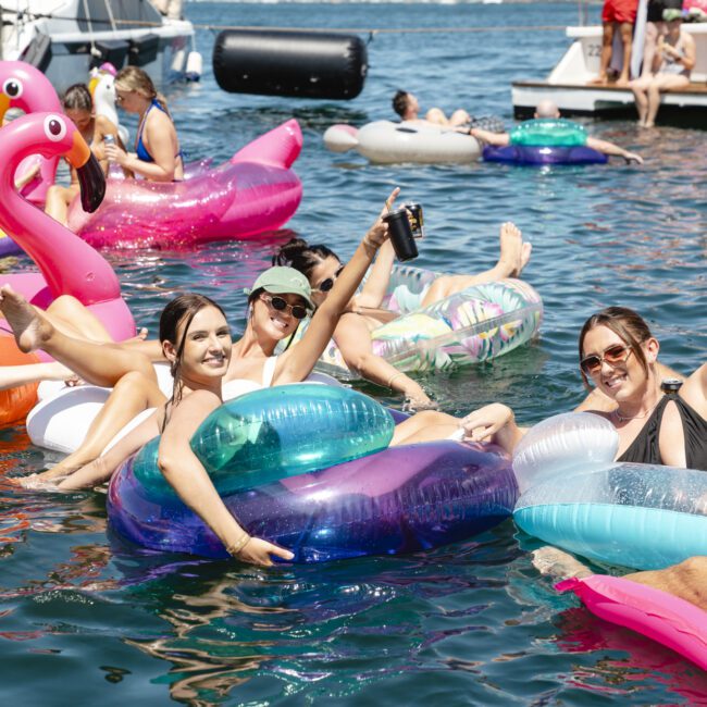 A group of people enjoying a sunny day on a lake, sitting on colorful inflatable pool floats, including a flamingo and round tubes. They are smiling, holding drinks, and surrounded by more floaters with boats in the background.