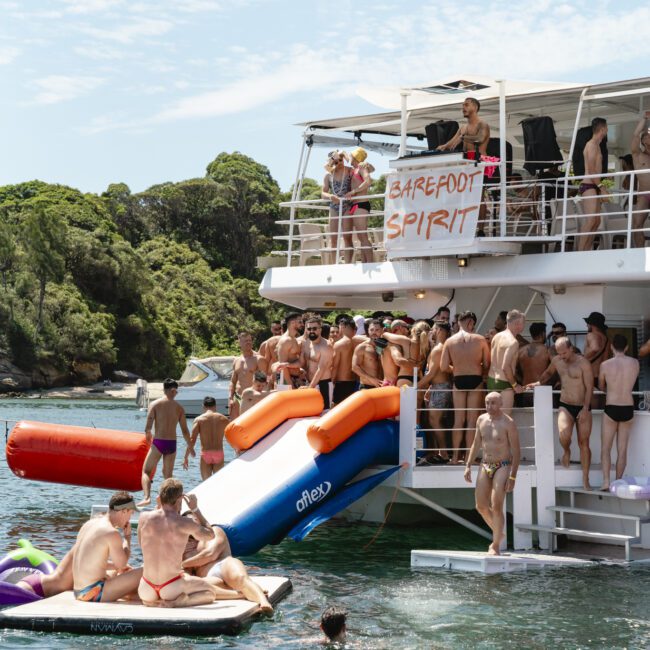 A group of people in swimwear are enjoying a party on a boat named "Barefoot Spirit." Some are on an inflatable slide leading to the water, while others are on a float nearby. The background features lush greenery and clear sky.