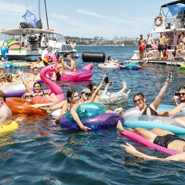 A group of people enjoying a sunny day on the water, floating on inflatable rings and loungers of various shapes and colors, including a pink flamingo. They are near anchored boats, with the city skyline visible in the distance.