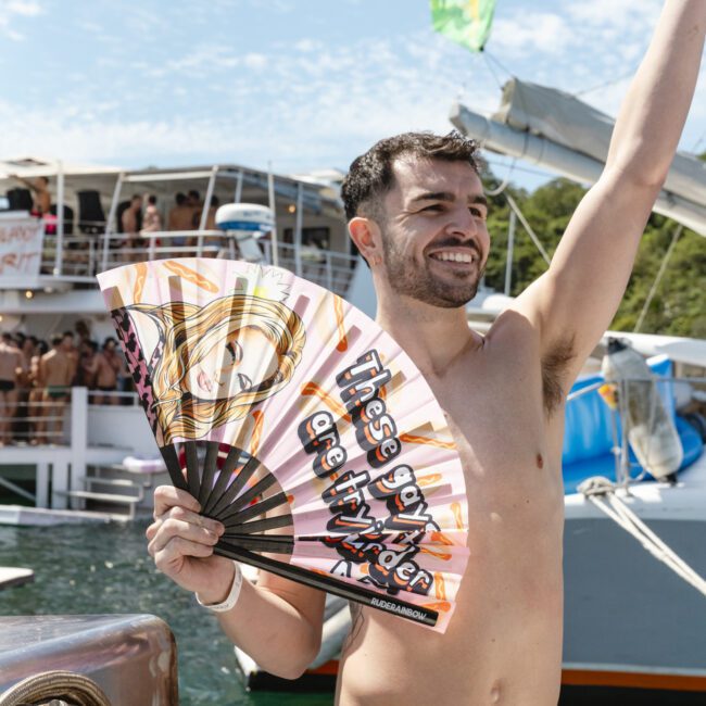 A smiling man in a colorful swimsuit holds a decorative fan with text on it, standing on a boat against a backdrop of other boats and people. The sky is clear and sunny.