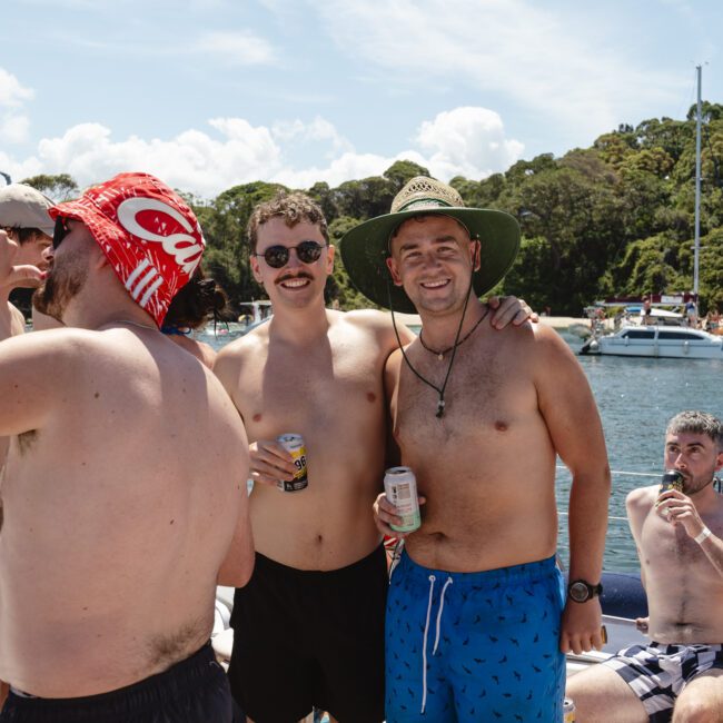 A group of men on a boat, enjoying a sunny day. Some are shirtless, holding drinks, and wearing hats or sunglasses. A forested shoreline and sailboats are visible in the background. The mood appears relaxed and cheerful.