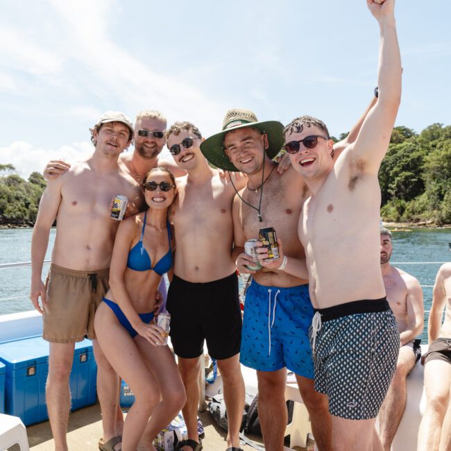 A group of six people in swimwear are posing happily on a boat in sunny weather. They are holding drinks, and one person is wearing a hat. There's a cooler and a lush green shoreline in the background.