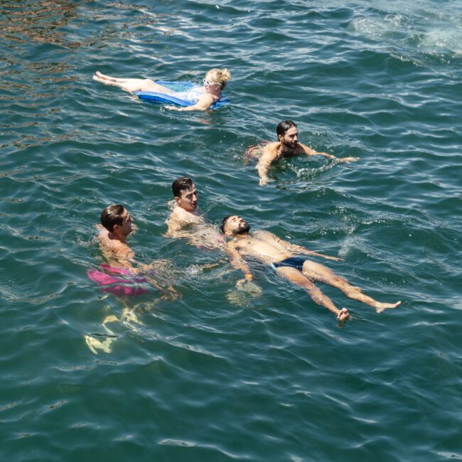 A group of people swims in clear, green water. One person is floating on their back, while others swim nearby. The scene is bright and sunny, creating a relaxing atmosphere.