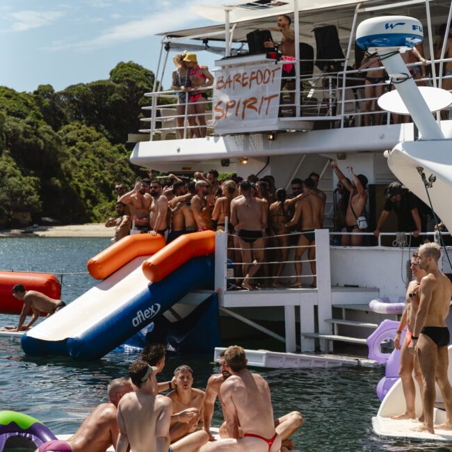 A lively group of people enjoys a day on a houseboat named "Barefoot Spirit." Some are standing on the deck, while others relax on floating platforms and inflatables in the water. A waterslide extends from the boat into the calm sea.