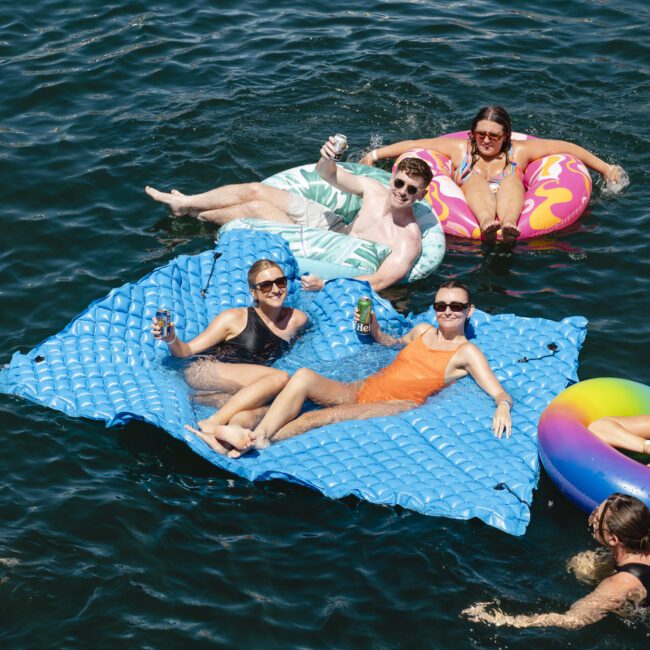 A group of people relax and enjoy drinks while floating on colorful inflatables in a sunny lake. They smile and raise their cans, surrounded by clear blue water. The scene is lively and cheerful, capturing a moment of leisure and fun.