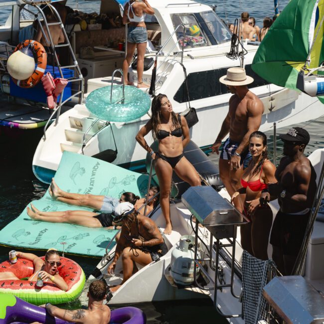 A lively scene of people enjoying a boat party on the water. They are sunbathing, lounging on inflatables, and socializing on and between several boats. Bright swimwear and sunny weather highlight the festive atmosphere.