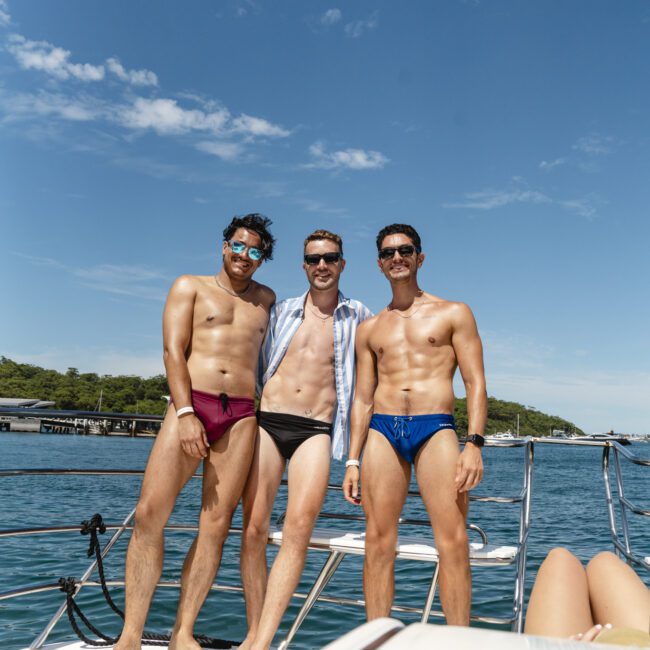 Three men smiling and posing on a boat in swimwear under a clear blue sky. One wears sunglasses, another sports a shirt, and water is visible in the background. A partial view of a person lounging is in the foreground.