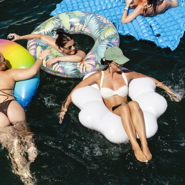 Three people relax on colorful pool floats in a body of water. One is on a rainbow float, another on a floral one, and a third on a white float. They enjoy the sun, wearing swimwear and sunglasses, some with beverages.