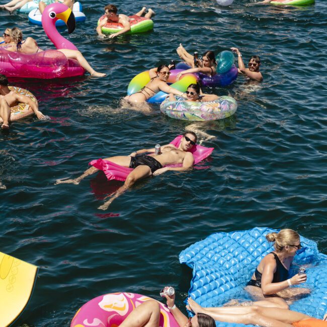 People are relaxing on colorful floaties in a lake. The group appears to be enjoying sunny weather, surrounded by inflatables like a flamingo and donut. Some people hold drinks, while others chat and sunbathe.