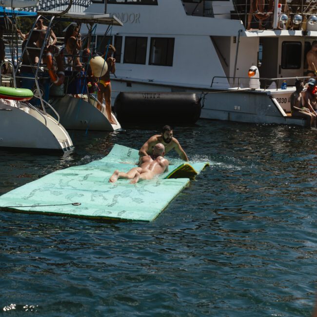 People enjoying a sunny day on a green floating mat in the water. Boats are anchored nearby with more people on board. The scene is lively, suggesting a fun outing by the water.