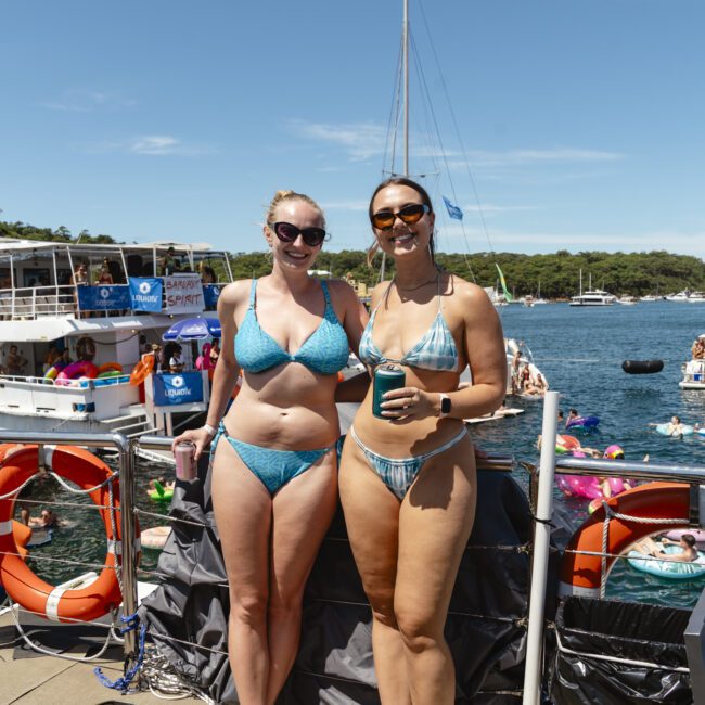 Two people in blue swimsuits stand on a dock by the water, smiling and holding drinks. Boats and colorful floaties are in the water behind them, under a clear blue sky.